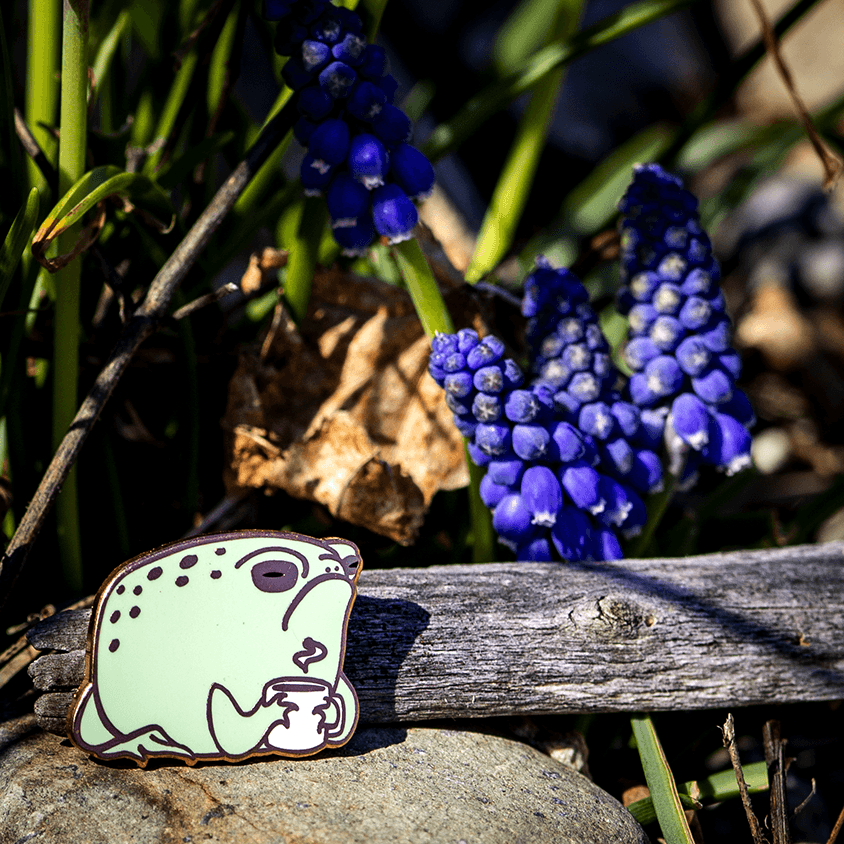 An enamel pin of Grumps the Desert Rain Frog holding his coffee mug by The Roving House