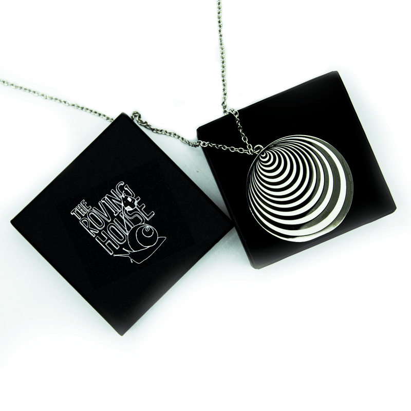 A large pendant of hypnotic black nickel and white enamel rings on a silver chain, draped over a black keepsake jewelry box. The box is printed with The Roving House snail logo in silver foil.