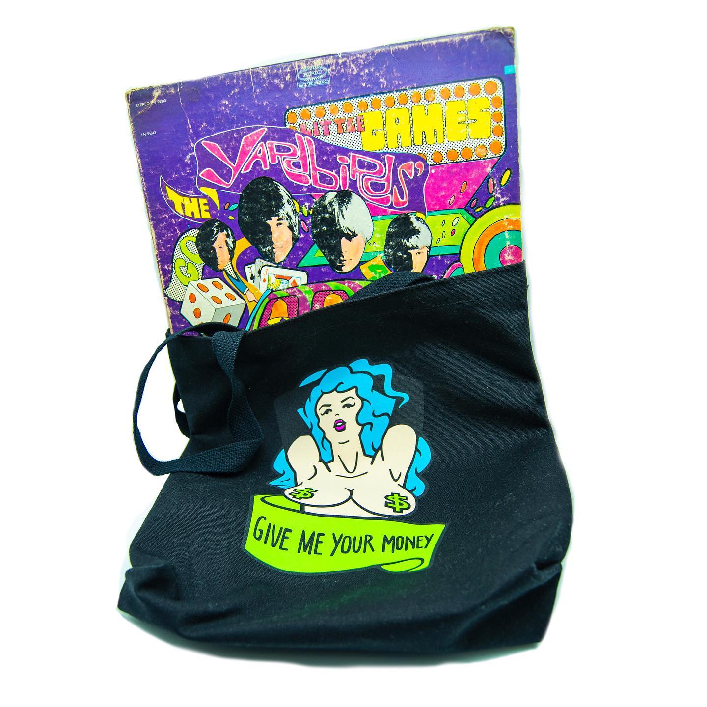 A black canvas tote bag, featuring the bust of a topless, blue haired woman wearing green dollar sign pasties. Below her is a banner that says "GIVE ME YOUR MONEY". The bag contains a Yardbirds record.