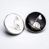 Two round enamel pins with The Roving House silver snail and bindle logo. One is white, and one is black.