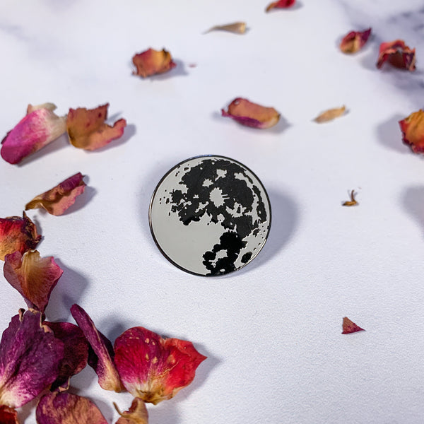 An enamel jewelry pin of a grey and nickel full moon, surrounded by red and pink rose petals.