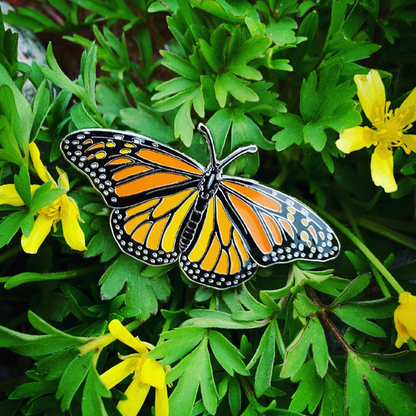 A black, orange, and white monarch butterfly enamel pin, displayed with yellow flowers.