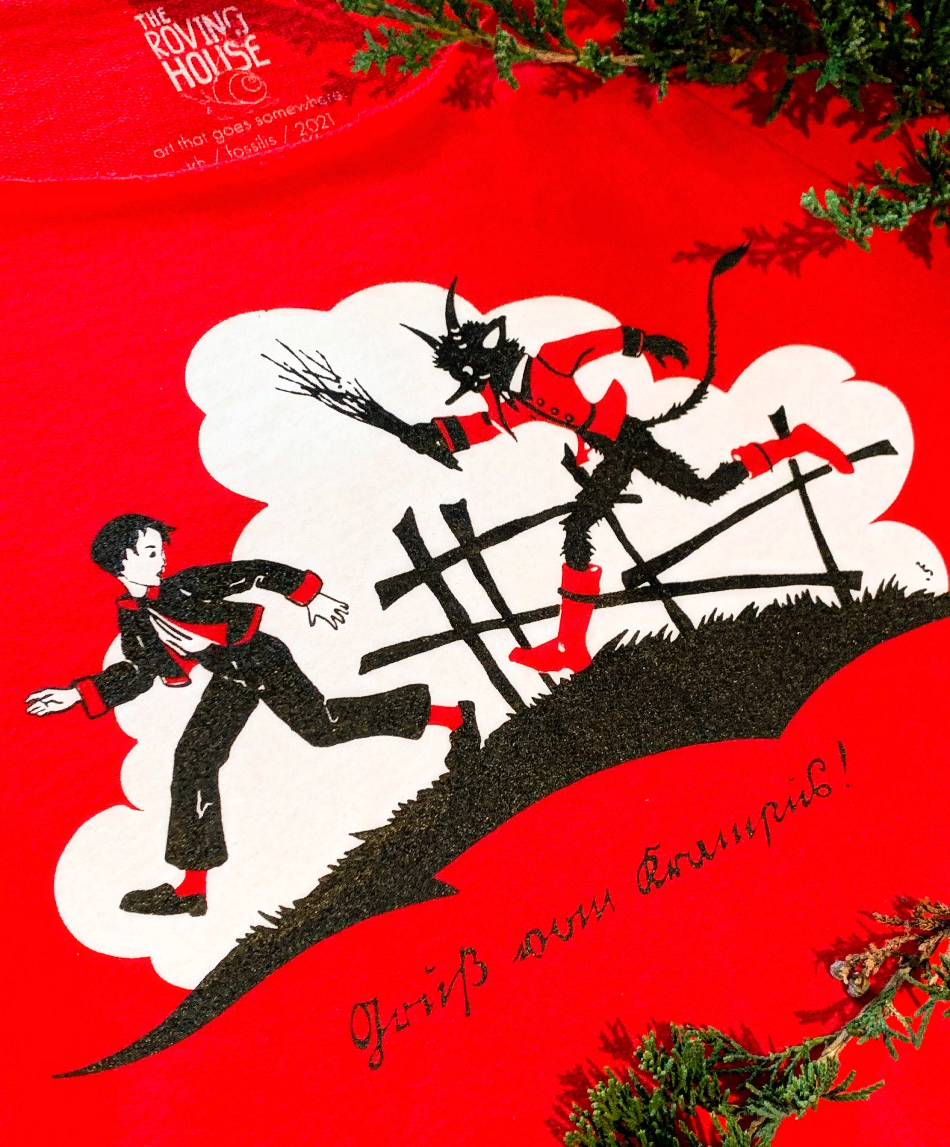 A closeup of a red, black, and white shirt design. Vintage 1900s artwork features Krampus with a bundle of sticks chasing a boy, with the words "Gruss vom Krampus!" (Greetings from Krampus!)