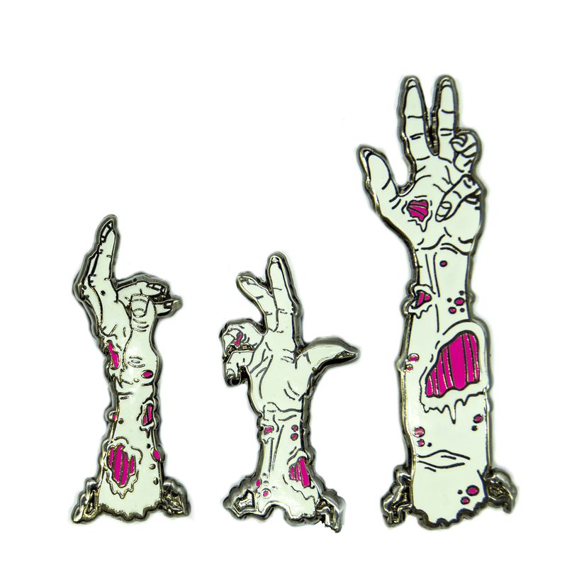 A set of three zombie arm pins, appearing to come up from the ground. Their skin is greenish white, and their flesh wounds are neon pink.