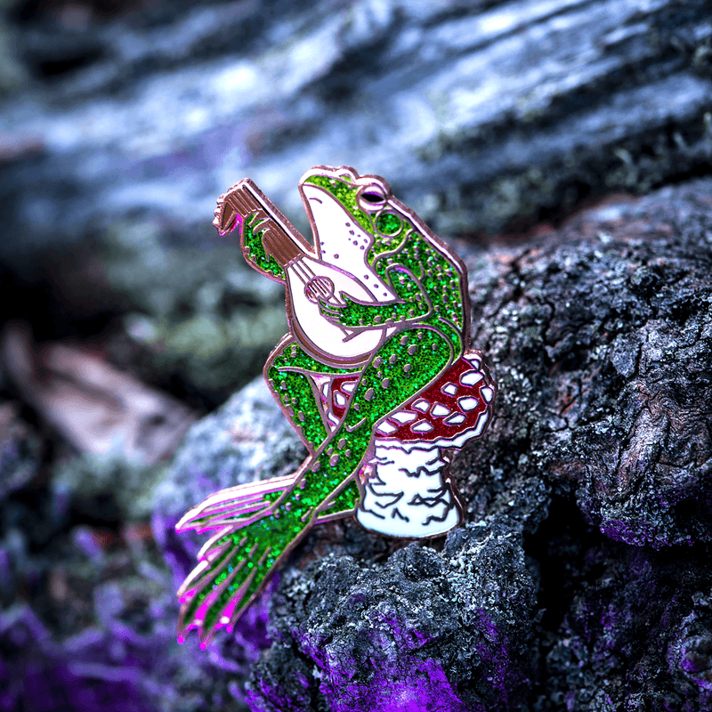 An enamel pin of a glittering green frog playing the lute, while sitting on a red fly agaric mushroom
