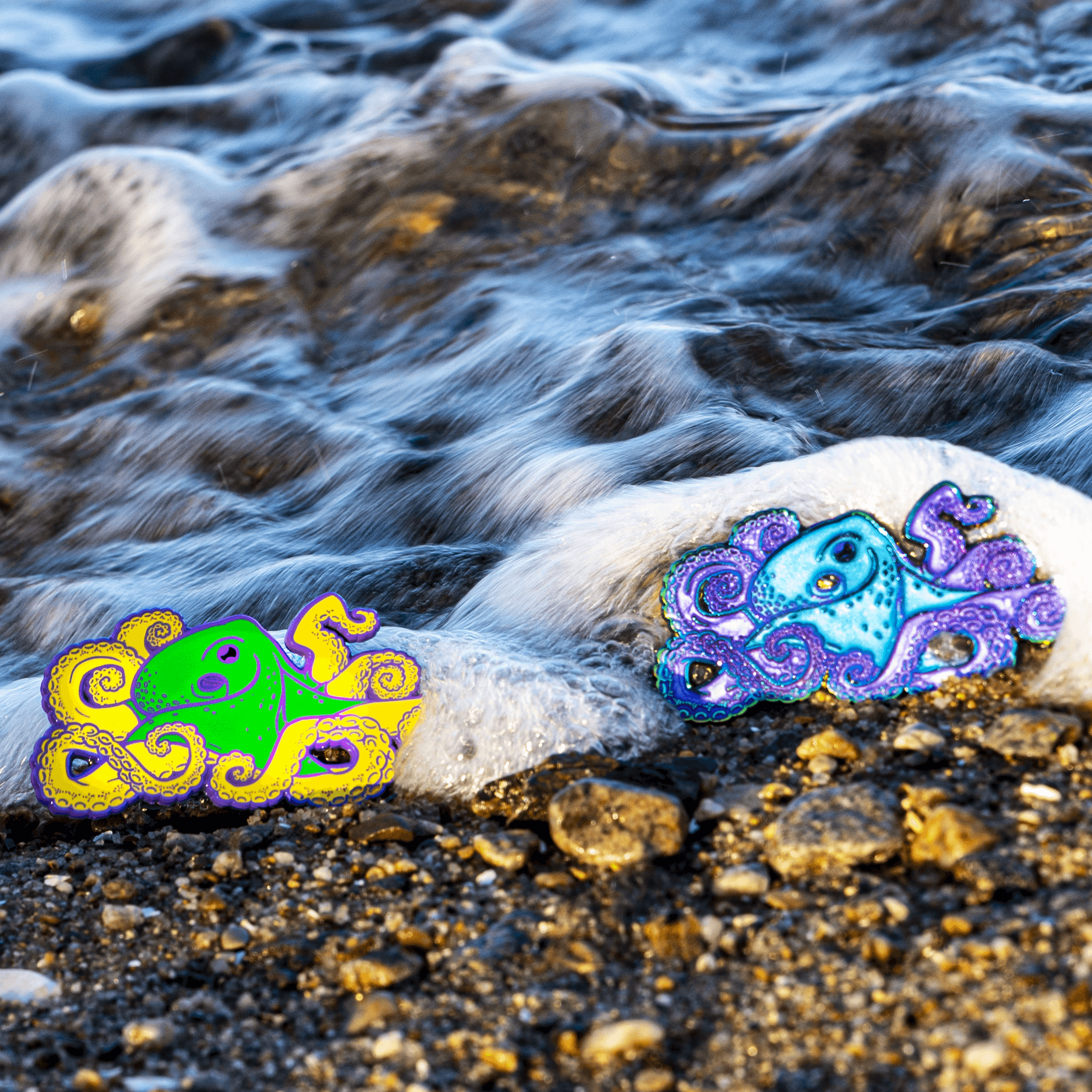 Two large octopus enamel pins sit on a pebbled beach, where they are splashed softly by the foamy sea.