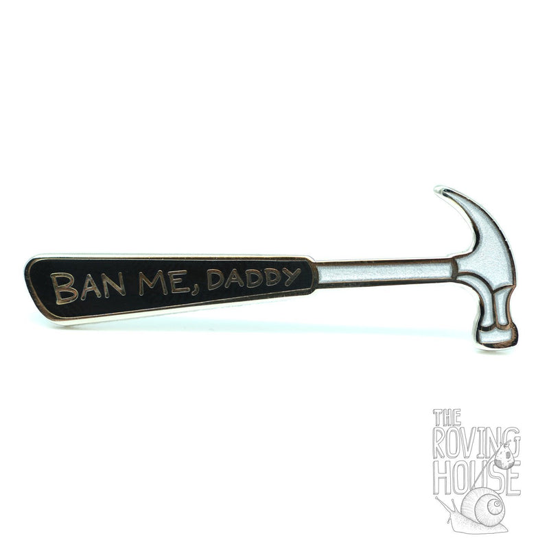 An enamel pin in the shape of a hammer, with "BAN ME, DADDY" written on the handle.