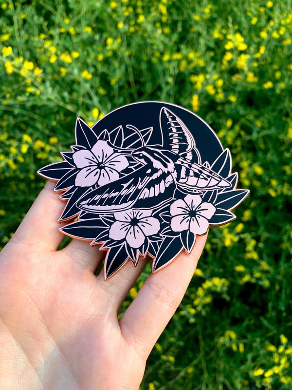 A woman's hand holding a large, rose gold and black enamel pin of a white lined sphinx moth hovering like a hummingbird over wild petunias. In the background are green and yellow flowers.