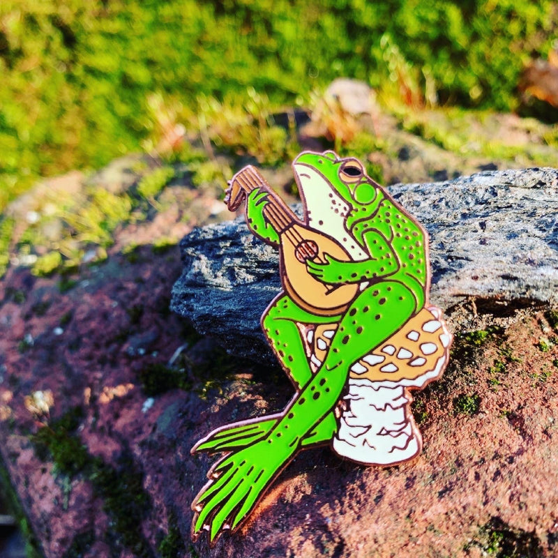 An enamel pin of a frog, playing a stringed instrument, sitting on a golden agaric mushroom. In the background is a charming setting of stone and moss.