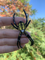 A woman's hand holding a life-sized black and yellow Argiope aurantia enamel pin.