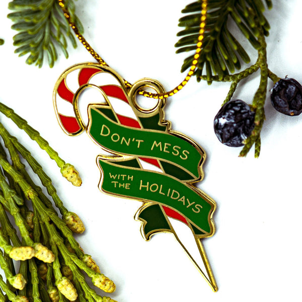 An enamel ornament of a red, white, and silver candy cane sharpened at one end, surrounded by a green banner that reads "DON'T MESS WITH THE HOLIDAYS". Evergreen branches and juniper berries surround the ornament.