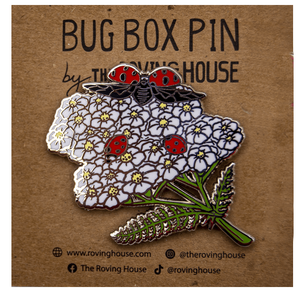 An enamel pin of three red and black nine-spotted ladybugs exploring white yarrow blossoms.