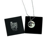 A grey enamel and black nickel pendant of the full moon on a silver metal chain, draped over a black jewelry box with The Roving House snail logo printed on its cover.