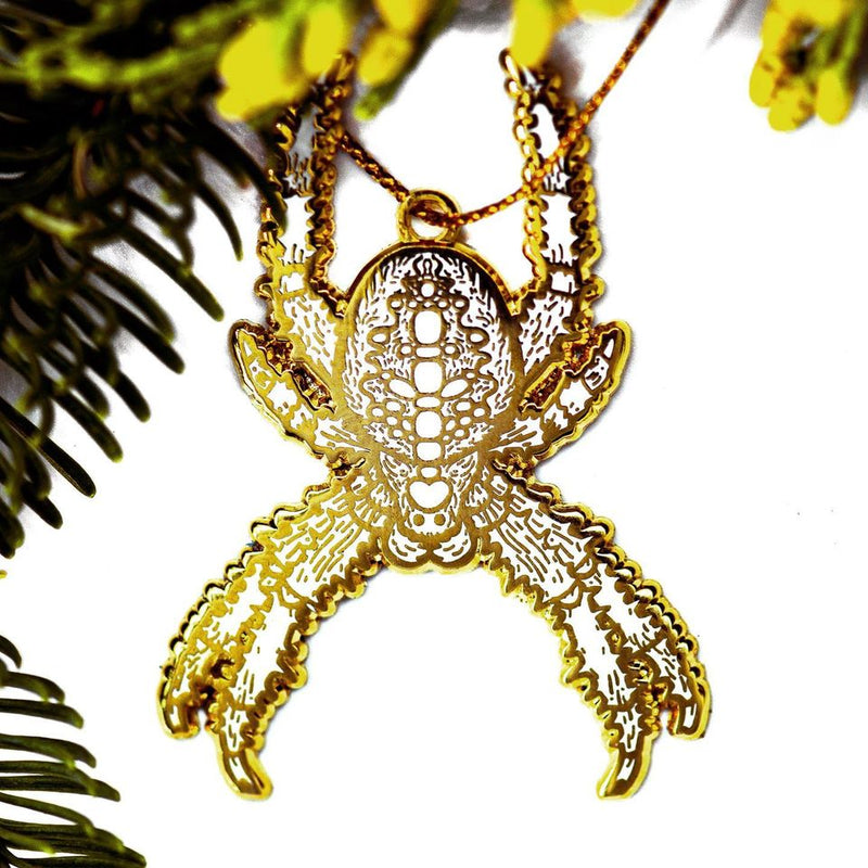 A gold and white enamel holiday tree ornament of a cross orb weaver Christmas Spider danging from a golden string on an evergreen bough. 